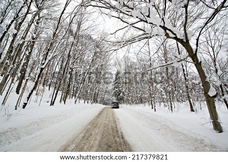 A car is parked down a rural road covered in snow with deep snow banks and snow covered trees on either side of the road during the winter season.
