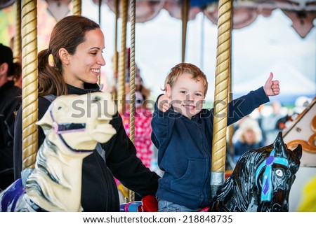 A happy mother and son are riding on a merry-go-round carousel together, smiling and having fun at a fair or amusement park.  The boy holds two thumbs up.