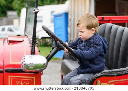 A happy young boy sits in an old shiny vintage red fire truck holding on to the steering wheel pretending to drive.