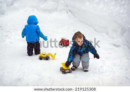Two boys are playing outside with toy trucks in the snow during a snowfall during the winter season.  There are deep snowbanks around them.