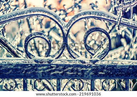A close up of part of an ornamental wrought iron gate with curve patterns covered in a thick layer of ice and icicles after an ice storm.  Filtered for a retro, vintage look.