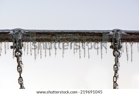 A close up of the metal bar and chains hanging down of a swing at a playground covered in thick ice and icicles after an ice storm.
