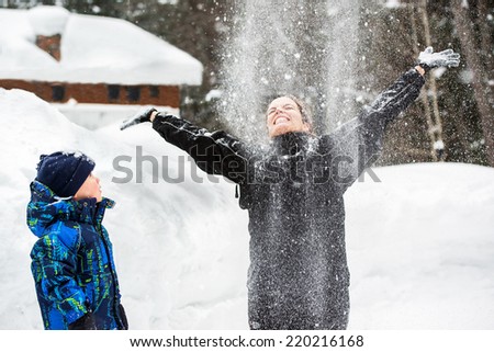 A young boy watches his mother throwing snow up in the air smiling with her arms out as it falls on her face.
