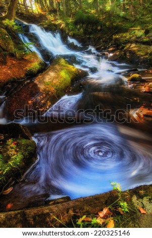 A waterfall in the background in a forest landscape with swirling pools of water in the foreground during the fall season.  A long exposure was used to veil the flowing motion of the water.
