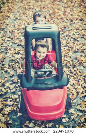 Playing outside, a young happy boy pushes his younger brother who is smiling with joy in a toy car on a driveway covered in fall leaves during the fall season.  Filtered for a retro, vintage look.
