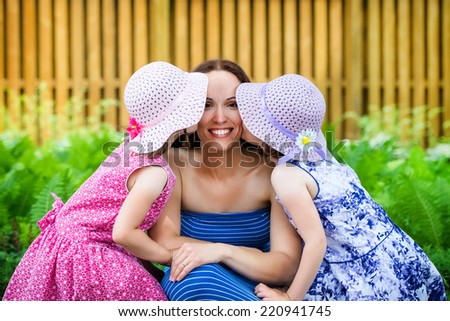 A family picture of two daughters on either side of their mother each giving a kiss on her cheek outside on a sunny spring day.