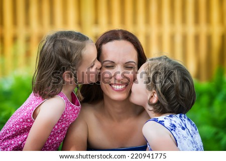 A close up family picture of two daughters on either side of their mother each giving a kiss on her cheek.