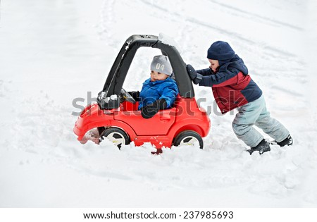 A young boy dressed for cold weather sits in a red toy car stuck in the snow during the winter season.  His older brother helps by giving the car a push from behind.