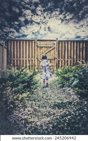 A little girl walks down a path toward a wooden gate in a fenced in garden looking for Easter eggs during an Easter egg hunt.  There are clouds in a darkened sky above.  Room for copy space.