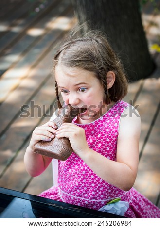 A little girl in a pink dress with wide eyes sits outside taking a big bite out of a large chocolate bunny on Easter day during the spring season.  Part of a series.