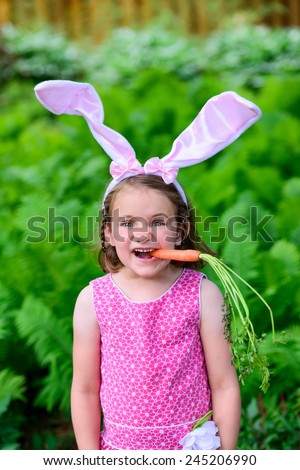 A young girl wearing Easter bunny ears making an expression holds a fresh carrot in her mouth outside in a lush garden setting during the spring season.   Part of a series.