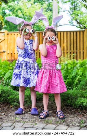 A funny portrait of two girls smiling and laughing, having fun on Easter wearing bunny ears and holding up silly eyes made from eggs outside in a garden during the spring season.  Part of a series.