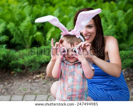 Funny photo of a smiling little boy having fun on Easter wearing bunny ears outside in a garden during the spring season.  His mother holds up silly eyes for him made from eggs.  Part of a series.