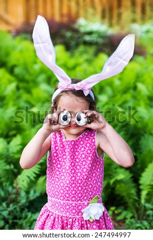 A funny portrait of a little girl having fun on Easter wearing bunny ears and holding up silly eyes made from eggs outside in a garden during the spring season.  Part of a series.