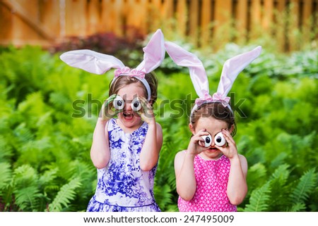 A funny portrait of two girls having fun on Easter wearing bunny ears and holding up silly eyes made from eggs outside in a garden during the spring season.  Part of a series.