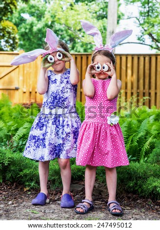 A funny portrait of two girls having fun on Easter wearing bunny ears and holding up silly eyes made from eggs outside in a garden during the spring season.  Part of a series.