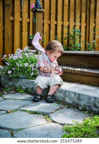 Boy with bunny ears resting on his shoulders spends a quiet moment for himself to sit on a step and peel an egg he finds during an Easter hunt in the spring season in a beautiful garden setting.