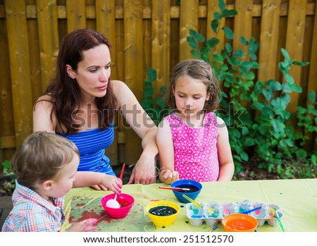 Family painting and decorating eggs.  Mother looks on at her children having fun as they color dye their Easter eggs outside during the spring season in a garden setting.  Part of a series.