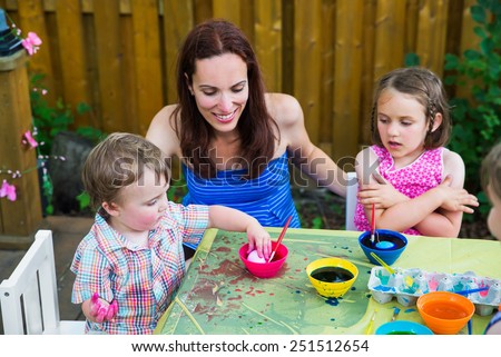 A Mother smiling at her boy decorating eggs as he puts his Easter egg in pink color dye while outside  During the spring season in garden setting.  Sister watches.  Part of a series.