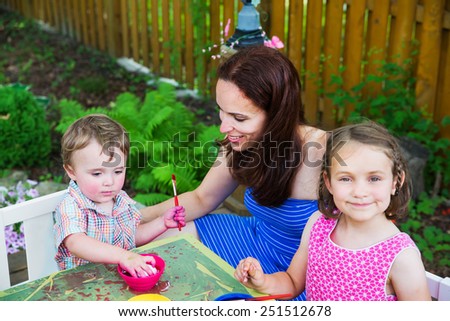 Family painting and decorating eggs outside during spring season in a garden setting.  Mother smiles at her boy color dying an Easter egg pink   A girl  in the foreground smiles.  Part of a series.