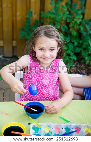 A happy girl sits outside in a springtime garden setting at a crafts table painting and decorating Easter eggs.  she smiles proudly and holds up a freshly colored blue dyed egg.   Part of a series.