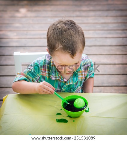 A little boy sitting at a crafts table having fun painting and decorating Easter eggs.  He colors an egg in a bowl with green dye while outdoors in the spring season.  Part of a series.