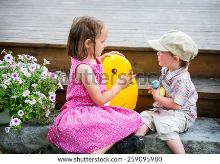 A boy holding on to colorful Easter eggs plays with a girl sitting beside him holding a toy chick outside during the spring.  Part of a series.