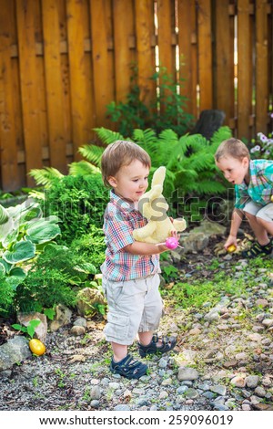 A boy plays with a plush toy bunny rabbit while his big brother looks for Easter eggs during an egg hunt outdoors in a garden.  Part of a series.
