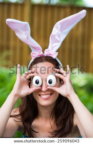 A portrait of a woman smiling wearing Easter rabbit ears holding up silly eyes made from eggs in front of her eyes outside in a garden during the spring season.  Part of a series.