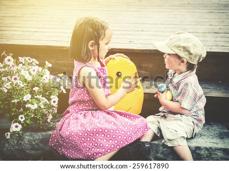 A boy holding on to colorful Easter eggs plays with a girl sitting beside him holding a toy chick outside during the spring.  Part of a series.  Filtered for a retro, vintage look.