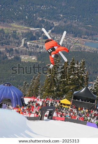 A skier in a bright orange suit flips upside down during a ski halfpipe competition.