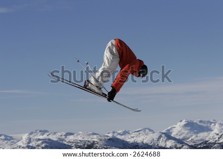 A skier doing a truck driver air spins high off a jump over a view of mountains in the background.