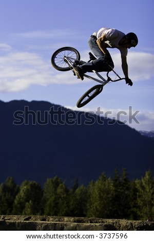 A BMX bike rider performs an aerial jump trick off a dirt ramp.