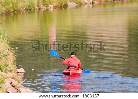 A kayaker in a bright red craft, with an equally bright red jacket paddles along the edge of a quiet lake.
