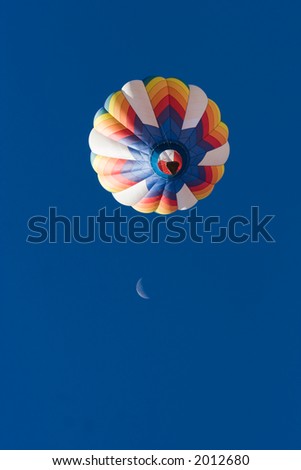 A brightly colored hot air balloon floats in the blue New Mexico sky with a quarter moon in the background