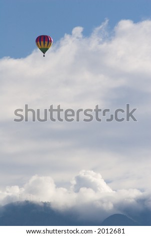 A brightly-colored hot air balloon floats in the blue New Mexico sky with cloud-covered Sandia Mountains in the background - vertical orientation
