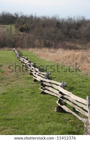 A split-rail fence runs down a hill next to a field, to a streambed, then up a hill in the distance