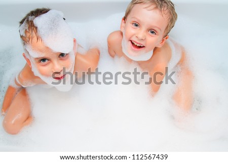 Two little boys - brothers in white bath tub covered with soap foam having bath