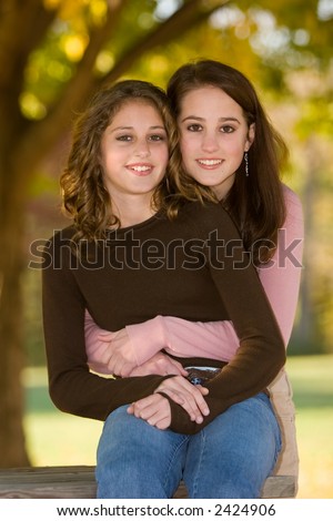 16 year old big sister hugging 12 year old little sister outdoors under tree in early fall.