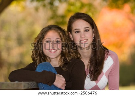 16 year old big sister beside 12 year old little sister outdoors under trees in early fall.