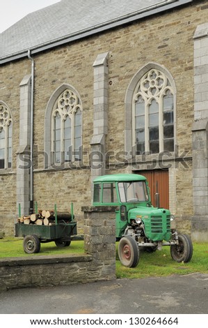 old tractor and church, ardennes, foreshortening of old tractor with cart carrying wood,  in front of church of agricultural village, shot in typical cloudy light