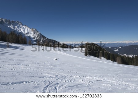 snowy meadows under Latemar, Costalunga pass; bright snow over meadows on smooth slopes in Dolomites under famous mountain range, in distance Bolzano valley under deep blue sky