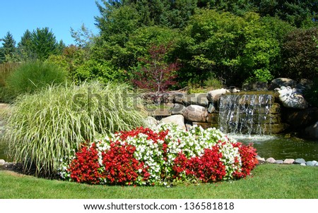 Landscape background with red and white mixed flower beds blossoming against green tall grass with a waterfall background