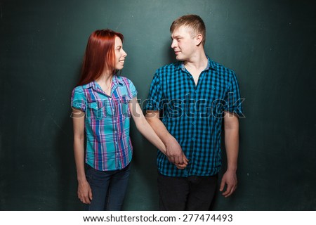 Beautiful Young Couple Over Dark Green Wall. They look in eyes each other and smile. Man takes his girlfriend by hand and leads. Love story. Red-haired woman and blonde man in check shirts.