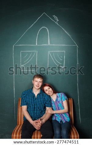 Beautiful Young Couple in house drawn with chalk. Affordable housing for young families, mortgage. Man and woman sit on a sofa having embraced. Red-haired woman and blonde man in check shirts.