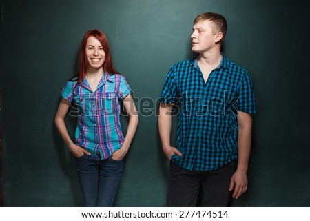 Beautiful Young Couple Over Dark Green Wall. Man looks at woman with interest. Love story, acquaintance, flirtation. Red-haired woman and blonde man in check shirts.