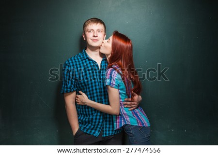 Portrait Of Beautiful Young Couple Over Dark Green Wall. Girl kisses her boyfriend gently. Love story. Red-haired woman and blonde man in check shirts.