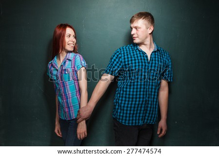 Beautiful Young Couple Over Dark Green Wall. They look in eyes each other and smile.  Man takes his girlfriend by hand and leads. Love story. Red-haired woman and blonde man in check shirts.