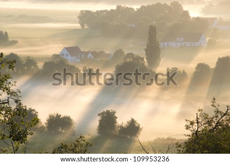 Rays of morning light illuminate fields and houses in the Netherlands on a foggy morning.