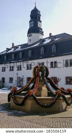 On a marketplace in Thuringia in Germany is at Easter decorated the fountain with many colorful Easter eggs, in the background a large house with tower/ Easter in Thuringia